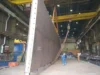 Workers guiding a large vertical steel plate being lifted by a crane in an industrial workshop.