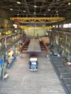 A large steel structure being lifted by a crane inside an industrial warehouse, with a truck positioned below.
