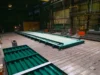 A large green metal structure laid out on the floor inside an industrial warehouse, with various equipment and tools in the background.