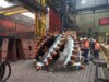A large industrial component being held in place by a crane inside a factory, surrounded by workers in orange safety vests and helmets.
