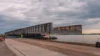 A large steel structure being transported on a heavy-duty vehicle along a riverside under a cloudy sky.