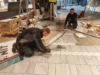 Two workers installing ceramic tiles on a floor, with one worker aligning tiles and the other preparing tools in a construction environment.