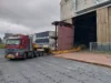 A large piece of a ship’s hull being transported by a truck from a shipyard building.