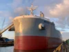 Large cargo ship named docked at a shipyard, with a clear view of its bow and anchor under a partly cloudy sky.