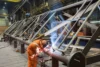 Industrial welder in orange protective gear welding a large metal framework inside a factory setting