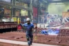 A worker in protective gear guiding a heavy chain inside a large industrial facility, with another worker welding metal beams in the background.