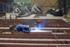 A worker in protective gear welding metal beams on a large factory floor, with heavy industrial machinery and large circular parts in the background.