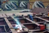 Two workers in protective gear welding large metal components in a factory, surrounded by industrial equipment and large circular metal parts in the background.