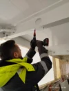 A worker using a power drill to secure a bolt into a ceiling structure inside a building, wearing a high-visibility jacket and gloves.