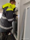 A worker in a high-visibility jacket using a power drill to work on a metal frame near a window inside a building.
