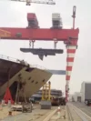 A shipyard scene where a large section of a ship is being lifted by a massive red crane, with the bow of a ship under construction visible in the foreground.