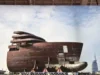 Partially constructed ship hull on a transport platform, with a modern building in the background under a cloudy sky.