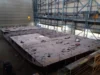 Large flat section of a ship's hull under construction in a shipyard, with scaffolding, equipment, and overhead cranes visible.
