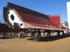 A large, partially constructed ship hull section with a red and white exterior, supported on temporary stands in an outdoor shipyard.
