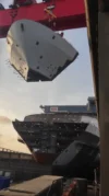 Large ship section being lifted by a red crane with another partially constructed ship hull visible in the background.