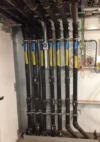 Section of an industrial facility showing multiple black pipes with yellow labels, neatly aligned and running vertically along a wall.