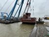 Large crane lowering a partially constructed vessel into the water at a shipyard.