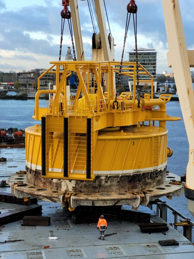 Large yellow industrial structure, part of an offshore equipment, with a waterfront and buildings in the background.