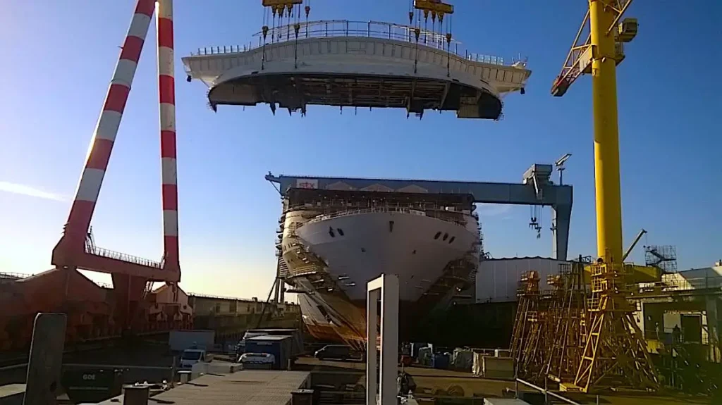 Ship under construction in a shipyard, with a large section being lifted by cranes.