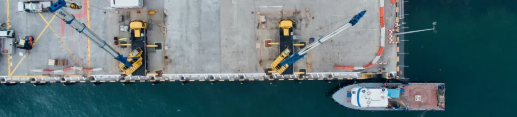Aerial view of a dock with cranes, vehicles, and a moored boat, showing the operations and layout of the port area.