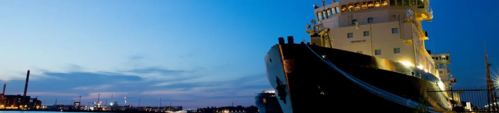 Large cargo ship docked at a port during twilight, with city lights and industrial buildings visible in the background.