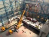 Aerial view of an industrial warehouse with a large yellow crane lifting a heavy load onto a platform, surrounded by other machinery and workers.