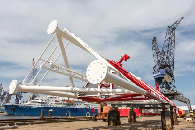 Large white and red steel structure at a shipyard, with a crane and docked ship in the background.