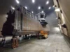 Large ship section under construction in a shipyard, with scaffolding, equipment, and overhead lighting illuminating the structure.