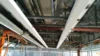 Ceiling view showing exposed pipes, electrical wiring, and support beams in an unfinished industrial space.
